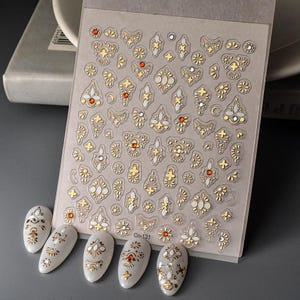 Puede incluir: Hoja de pegatinas de arte de u&ntilde;as con dise&ntilde;os dorados, blancos y anaranjados con forma de joya. La hoja incluye varias formas, incluidos patrones florales y geom&eacute;tricos. Se muestran cinco u&ntilde;as pintadas con dise&ntilde;os a juego debajo. El texto "Co-131" es visible.