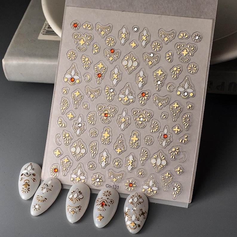 Puede incluir: Hoja de pegatinas de arte de u&ntilde;as con dise&ntilde;os dorados, blancos y anaranjados con forma de joya. La hoja incluye varias formas, incluidos patrones florales y geom&eacute;tricos. Se muestran cinco u&ntilde;as pintadas con dise&ntilde;os a juego debajo. El texto "Co-131" es visible.