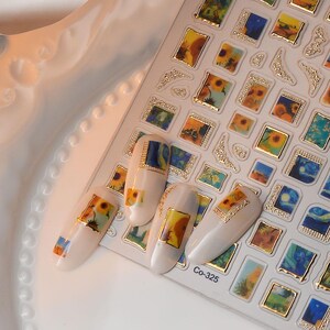 May include: Nail art stickers featuring miniature paintings of sunflowers and landscapes. The stickers have gold frames and are applied to white nail polish. The image includes several manicured nails with the stickers applied, showcasing the design.