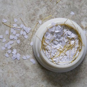 May include: A white container filled with gold glitter and white confetti. The container is on a light gray surface with white confetti scattered around it.