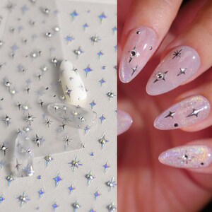 May include: A sheet of nail art stickers with holographic star designs. The stickers are shown applied to a set of nude-colored nails.