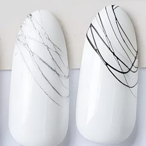 Puede incluir: Cuatro muestras de esmalte de uñas de forma ovalada con líneas de diferentes colores sobre un fondo blanco. Las líneas son blancas sobre negro, plateadas sobre blanco, negras sobre blanco y doradas sobre blanco.