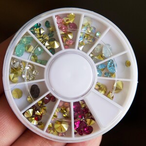 May include: A white plastic wheel with 12 compartments filled with various colored rhinestones. The rhinestones are in shades of green, blue, pink, and gold.