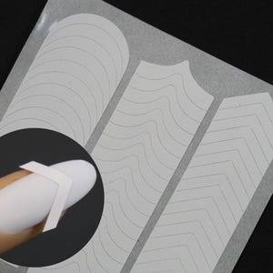 Pode incluir: Adesivos brancos para nail art com três designs diferentes: linhas curvas, linhas onduladas e padrões chevron. Os adesivos são mostrados em um fundo branco e em uma unha.