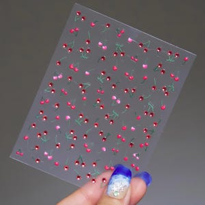 May include: Clear sheet of nail art stickers featuring a pattern of red and pink cherries with green stems and leaves. The cherries have a sparkling, gem-like appearance. The stickers are held by a hand with blue and white nail polish.