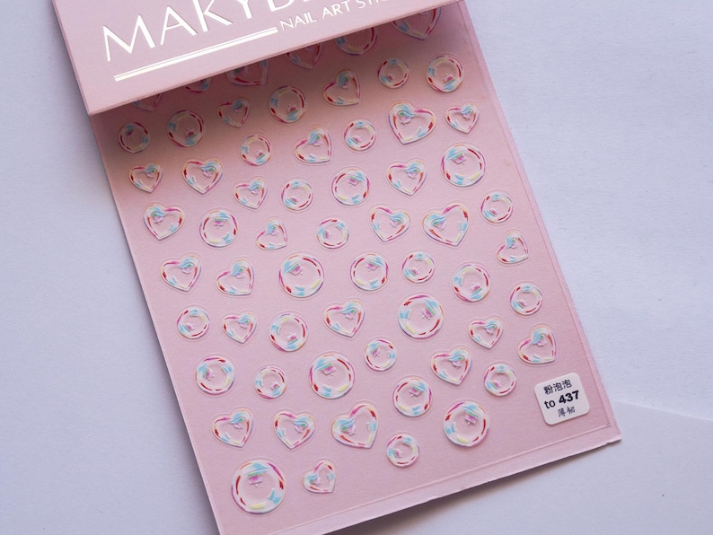 May include: Pink nail art sticker sheet with heart and bubble designs. The stickers have a clear, iridescent appearance with pink, blue, and white accents. The sheet includes the text "NAIL ART STICKERS" and a small label with Chinese characters.