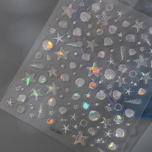 Peut inclure: Feuille transparente d'autocollants iris&eacute;s pour nail art avec des motifs marins. Les autocollants repr&eacute;sentent des &eacute;toiles de mer, des coquillages et de petites &eacute;toiles. La feuille porte la mention "Co-387".