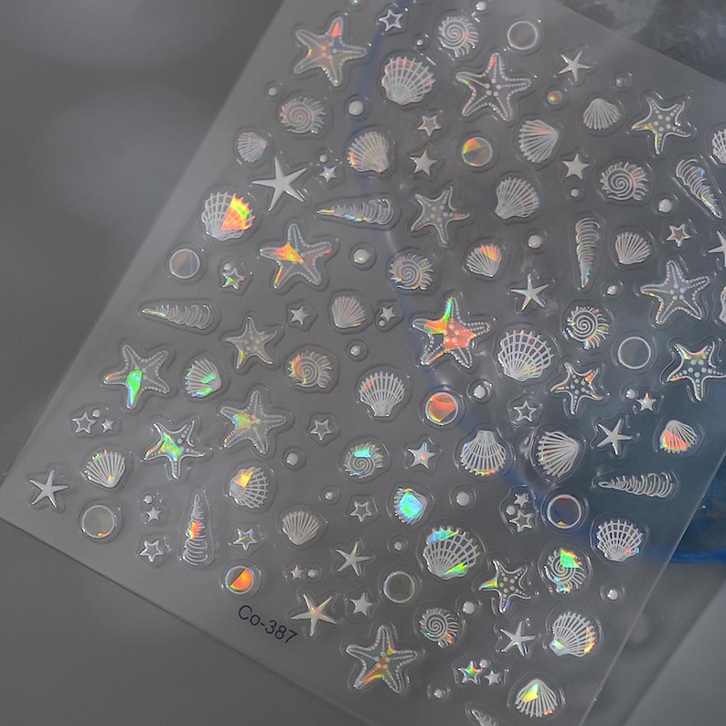 Peut inclure: Feuille transparente d'autocollants iris&eacute;s pour nail art avec des motifs marins. Les autocollants repr&eacute;sentent des &eacute;toiles de mer, des coquillages et de petites &eacute;toiles. La feuille porte la mention "Co-387".
