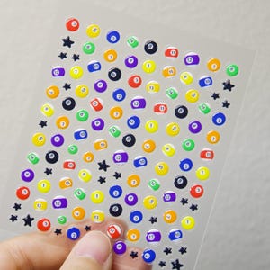 May include: A sheet of colorful, glossy stickers featuring billiard balls numbered 1 through 15, in various colors, and black star shapes. The stickers are on a clear, transparent background. The stickers are held by a hand.
