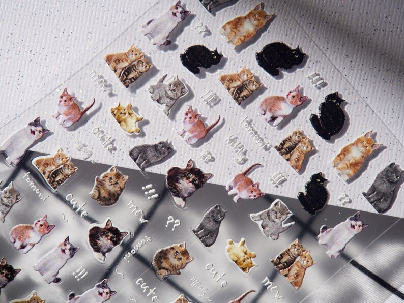 May include: Clear sheet of cat stickers in various colors and poses. The stickers feature different cat breeds, including black, gray, orange, and calico cats. Some stickers have the words "cute" and "meow" written on them.