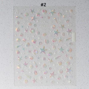 Peut inclure: Feuille transparente d'autocollants iris&eacute;s avec des formes sur le th&egrave;me marin. Les autocollants comprennent des &eacute;toiles de mer, des coquillages et d'autres motifs aquatiques. Le texte "#2" est visible en haut de la feuille. Les autocollants ont un effet holographique.