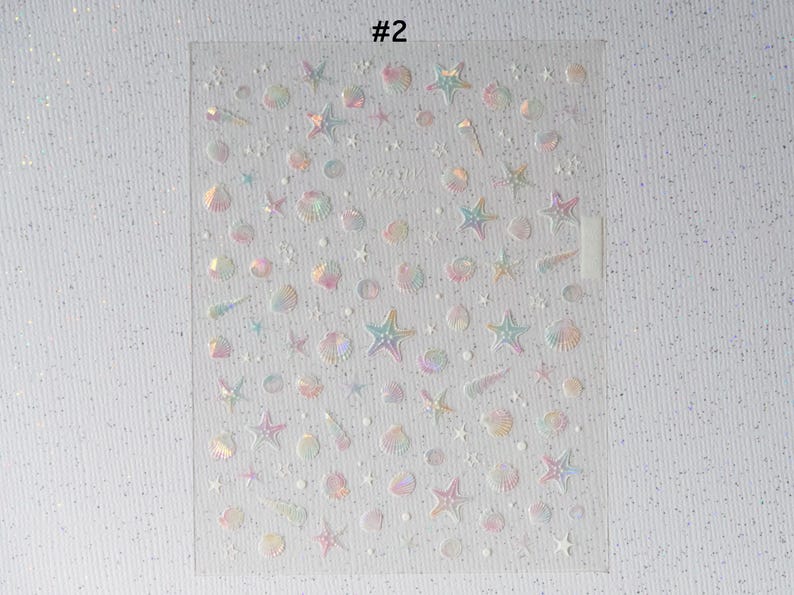 Peut inclure: Feuille transparente d'autocollants iris&eacute;s avec des formes sur le th&egrave;me marin. Les autocollants comprennent des &eacute;toiles de mer, des coquillages et d'autres motifs aquatiques. Le texte "#2" est visible en haut de la feuille. Les autocollants ont un effet holographique.