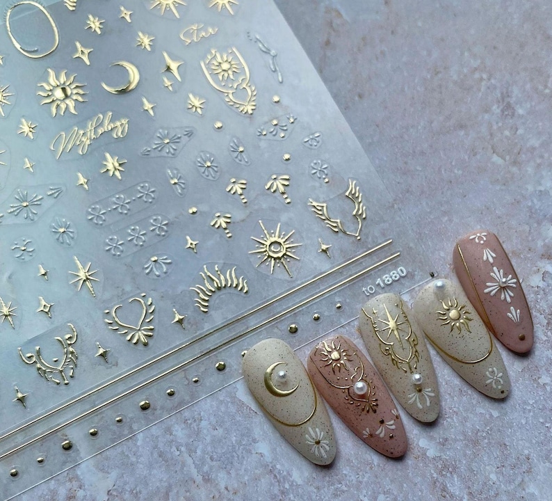 Pink and white nails with stickers on them in front of a sheet of gold celestial nail stickers