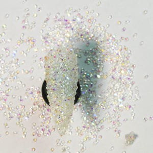 May include: A close-up of iridescent clear glass beads scattered on a white surface. The beads are small and round, with a slight sparkle. A small pile of the beads is gathered together in the center of the image.