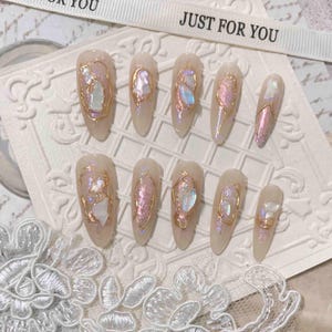 Może przedstawiać: Zestaw dziesięciu sztucznych paznokci z perłowym, tęczowym wykończeniem i złotymi akcentami z drutu. Paznokcie są długie i migdałowe. Paznokcie są ułożone w dwóch rzędach na białym tle.