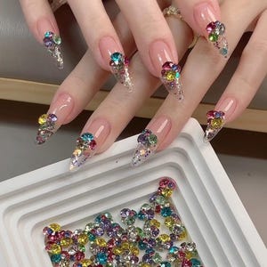 May include: Close-up of a hand with long, clear, stiletto nails decorated with colourful rhinestones. The nails are adorned with clusters of rhinestones in various shades of blue, green, red, yellow, and purple.