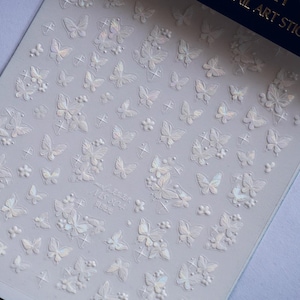 K&ouml;nnte beinhalten: Bogen mit Nagelkunst-Aufklebern mit einem sich wiederholenden Muster aus Schmetterlingen und floralen Designs. Die Aufkleber sind wei&szlig; mit schillernden Akzenten. Auf dem Bogen steht der Text "NG NAIL ART STICKERS".