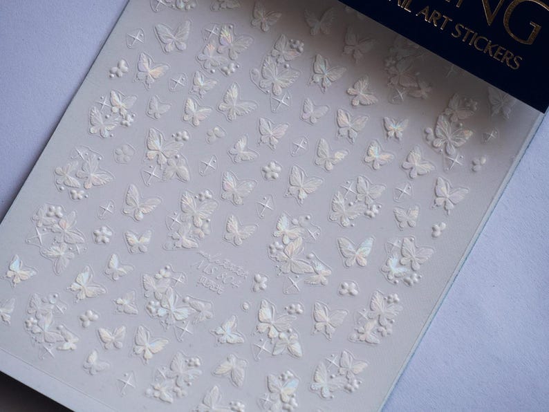 K&ouml;nnte beinhalten: Bogen mit Nagelkunst-Aufklebern mit einem sich wiederholenden Muster aus Schmetterlingen und floralen Designs. Die Aufkleber sind wei&szlig; mit schillernden Akzenten. Auf dem Bogen steht der Text "NG NAIL ART STICKERS".