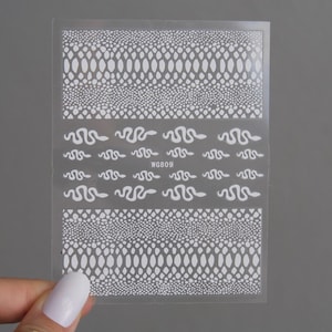 Puede incluir: Hoja de pegatinas transparentes para decoración de uñas con diseños de serpientes y escamas blancas. La hoja incluye diseños de serpientes y patrones de escamas repetidos. El texto "WG809" está impreso en la hoja.
