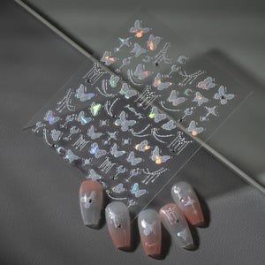 K&ouml;nnte beinhalten: Transparente Nagelkunst-Aufkleber mit Schmetterlings- und Himmelsmotiven. Die Aufkleber zeigen verschiedene Schmetterlingsformen, Monde, Sterne und andere dekorative Elemente. Mehrere manik&uuml;rte N&auml;gel werden mit den aufgebrachten Aufklebern gezeigt, die ein rosa-wei&szlig;es Farbverlaufsschema aufweisen.