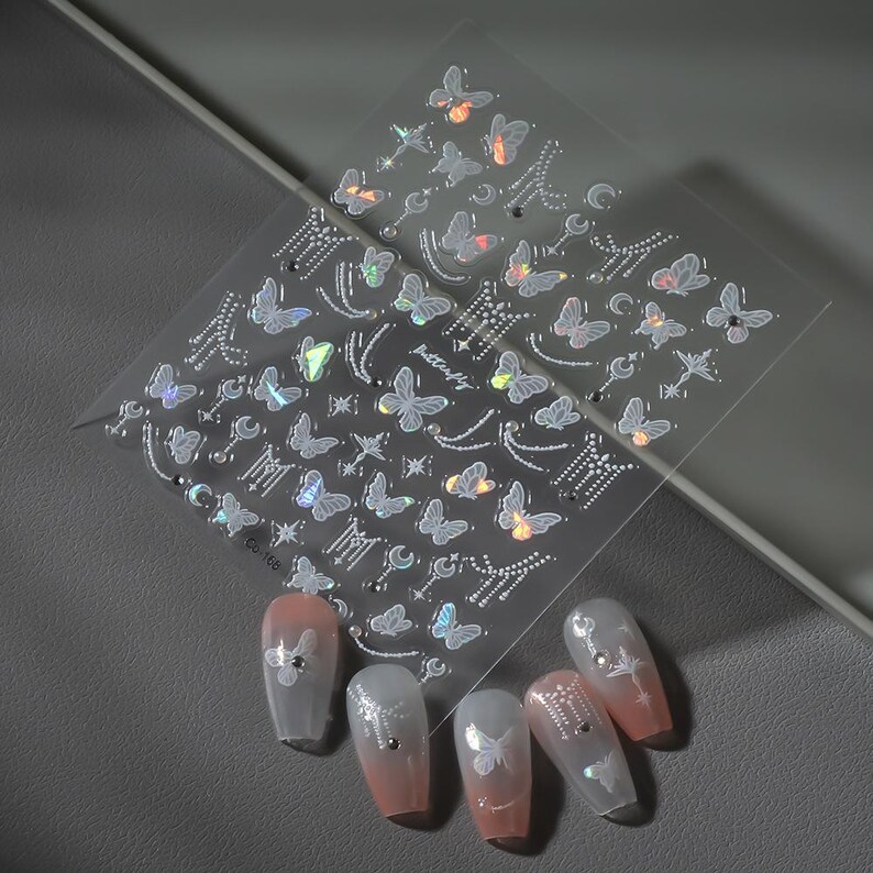 K&ouml;nnte beinhalten: Transparente Nagelkunst-Aufkleber mit Schmetterlings- und Himmelsmotiven. Die Aufkleber zeigen verschiedene Schmetterlingsformen, Monde, Sterne und andere dekorative Elemente. Mehrere manik&uuml;rte N&auml;gel werden mit den aufgebrachten Aufklebern gezeigt, die ein rosa-wei&szlig;es Farbverlaufsschema aufweisen.