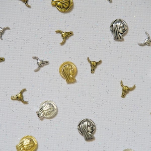 May include: An assortment of small, decorative metal charms in gold and silver tones. The charms include depictions of a woman's profile and bull horns, scattered on a white, glittery surface. These charms are likely for jewelry making or craft projects.