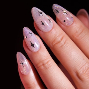May include: Close-up of a hand with five nails painted with a light pink glitter polish. Each nail has a black star design and some have small silver rhinestones.