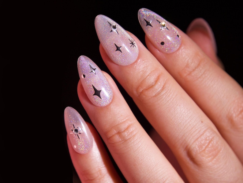 May include: Close-up of a hand with five nails painted with a light pink glitter polish. Each nail has a black star design and some have small silver rhinestones.