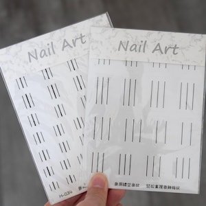 Puede incluir: Dos paquetes de pegatinas para uñas. Cada paquete es blanco con líneas verticales negras y las palabras "Nail Art" impresas en la parte superior. Los paquetes están en la mano de una persona.