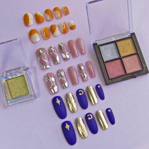 Puede incluir: Un conjunto de uñas postizas con diferentes diseños. Los diseños incluyen patrones abstractos en oro y plata, rosa y blanco, y azul y oro. También hay dos paletas de sombras de ojos con colores brillantes.