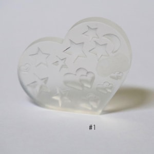 Puede incluir: Un sello de silicona transparente en forma de corazón con varios diseños, incluyendo estrellas, corazones y nubes. El sello está etiquetado como #1.