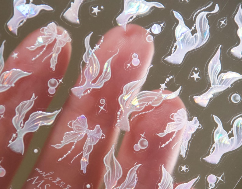 Peut inclure: Feuille d'autocollants transparente avec des motifs de queue de sir&egrave;ne, de n&oelig;uds et d'&eacute;toiles iris&eacute;s. Les autocollants ont une finition nacr&eacute;e et sont tenus par une main. Ces autocollants sont probablement destin&eacute;s &agrave; l'artisanat ou au nail art.