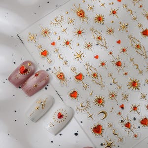 Peut inclure: Feuille transparente d'autocollants pour nail art avec des motifs célestes dorés, notamment des étoiles, des cœurs et des lunes. Plusieurs ongles peints sont présentés, avec des motifs assortis aux autocollants. Les ongles sont peints dans des tons de rose et de blanc.