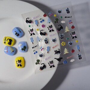 May include: A sheet of clear stickers with cartoon cat and nature-themed designs, including trees, cherries, and stars. The stickers are accompanied by several nail art designs in yellow and blue, featuring similar cat and tree graphics. The word "Miao!" is printed on the stickers.