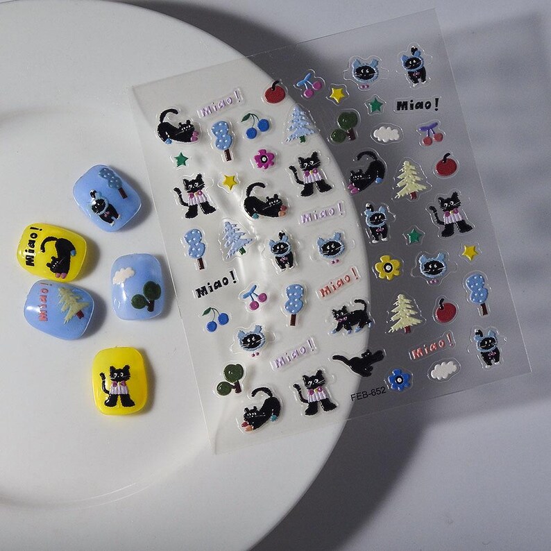 May include: A sheet of clear stickers with cartoon cat and nature-themed designs, including trees, cherries, and stars. The stickers are accompanied by several nail art designs in yellow and blue, featuring similar cat and tree graphics. The word "Miao!" is printed on the stickers.