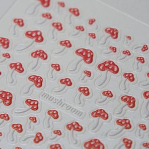 May include: White sheet with a repeating pattern of red and white mushroom stickers. The word "mushroom" is printed in a rectangular box. The mushrooms have red caps with white spots and white stems.