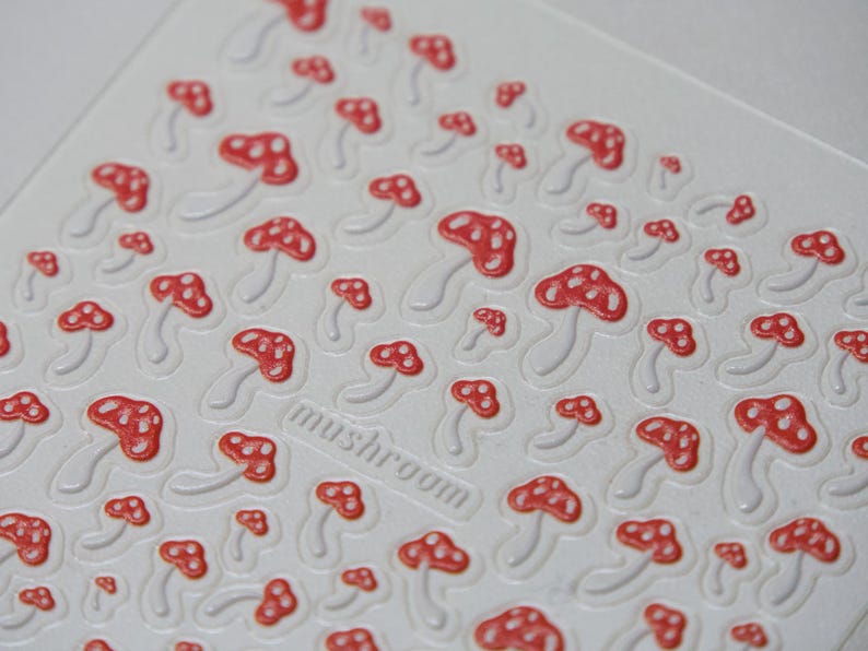 May include: White sheet with a repeating pattern of red and white mushroom stickers. The word "mushroom" is printed in a rectangular box. The mushrooms have red caps with white spots and white stems.