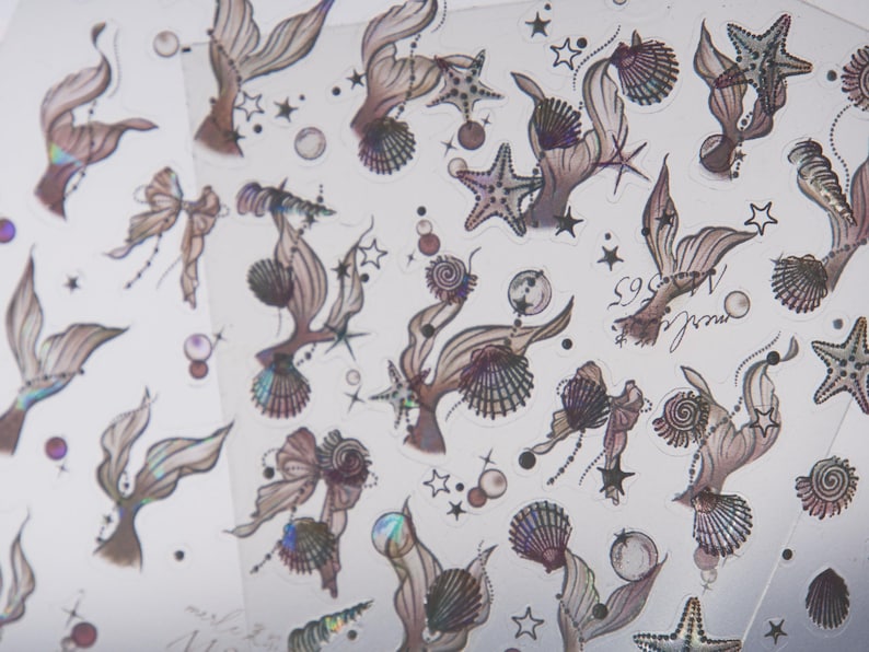 Peut inclure: Une feuille d'autocollants sur le th&egrave;me des sir&egrave;nes avec divers motifs. Les autocollants pr&eacute;sentent des queues de sir&egrave;nes, des coquillages, des &eacute;toiles de mer et des n&oelig;uds dans des tons de marron, de violet et de couleurs iris&eacute;es. De petites &eacute;toiles et bulles sont &eacute;galement incluses.