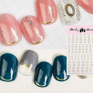 Op de afbeelding: Gouden nagelkunst stickers met een halve maan ontwerp. De stickers zijn op een wit vel met de tekst "Maky Nail" erop gedrukt. De stickers worden getoond op een set roze en teal nageltips.