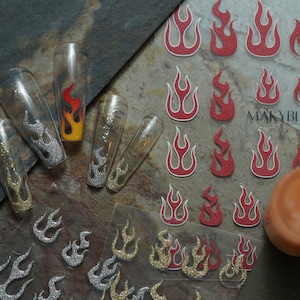 May include: A sheet of nail art stickers featuring flame designs in red, gold and silver. The stickers are on a clear background and are ready to be applied to nails. The sheet also includes the text "MAKYBLING".
