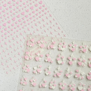 Op de afbeelding: Twee vellen met roze hart stickers. Eén vel heeft kleine roze harten op een witte achtergrond. Het andere vel heeft grotere roze en witte harten op een transparante achtergrond.