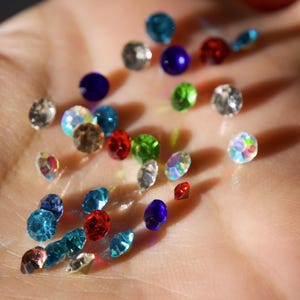 Op de afbeelding: Een verzameling kleurrijke strass-steentjes in verschillende vormen en maten, waaronder rond en druppelvormig. De edelstenen zijn in tinten blauw, rood, groen en iriserend, verspreid over een oppervlak.