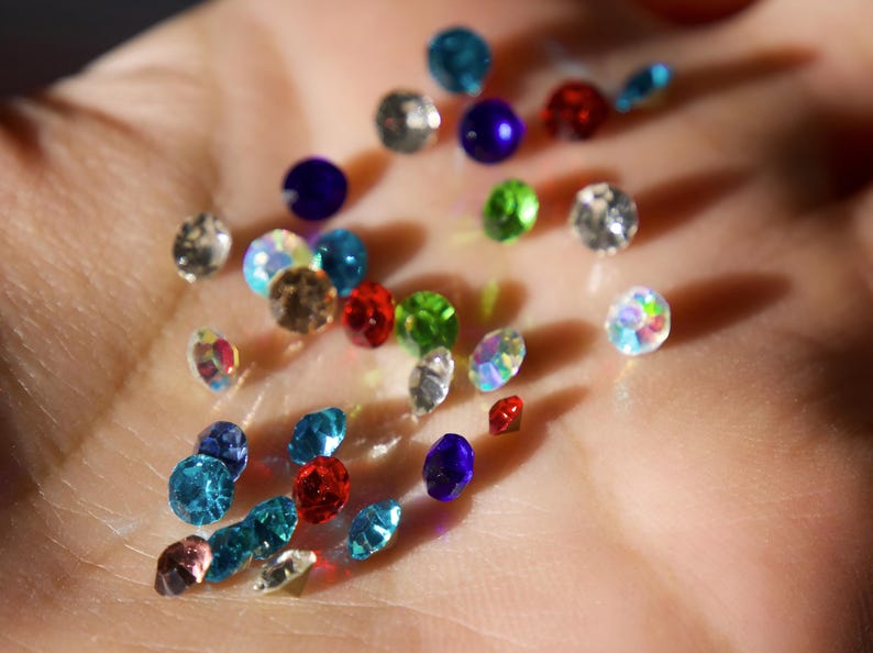 Op de afbeelding: Een verzameling kleurrijke strass-steentjes in verschillende vormen en maten, waaronder rond en druppelvormig. De edelstenen zijn in tinten blauw, rood, groen en iriserend, verspreid over een oppervlak.