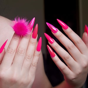 以下が含まれることがあります： 長く尖った明るいピンクの爪をした手のクローズアップ。いくつかの爪はキラキラした仕上げになっています。ピンクのふわふわのポンポンが1本の指にあります。