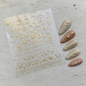 Peut inclure: Une feuille transparente d'autocollants pour ongles avec des motifs célestes en feuille d'or, incluant des soleils, des lunes, des étoiles et des formes géométriques. Cinq faux ongles en amande décorés sont présentés sur la droite.