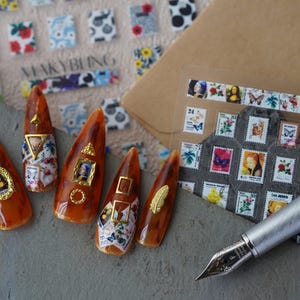 May include: Five brown nail tips with gold accents and miniature images of famous paintings. The nail tips are arranged on a grey surface with a silver pen and a sheet of decorative stickers with miniature images of stamps.