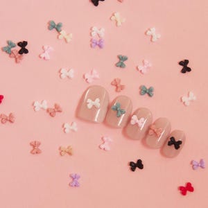 Könnte beinhalten: Eine Ansammlung kleiner, farbenfroher, schleifenförmiger Nagelkunstdekorationen, die auf einer rosa Oberfläche verstreut sind. Mehrere Nägel sind mit Schleifenakzenten verziert. Die Schleifen sind in verschiedenen Farben, darunter Weiß, Rosa und Schwarz.