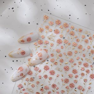 Può includere: Adesivi per nail art con disegni di rose dorate e rosa. Gli adesivi sono su un foglio trasparente, alcuni applicati su punte di unghie bianche. Le parole "Best Wishes" sono stampate.