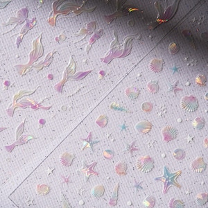 Peut inclure: Feuille d'autocollants iris&eacute;s avec des queues de sir&egrave;ne, des coquillages, des &eacute;toiles de mer et de petits cercles. Les autocollants ont une finition nacr&eacute;e avec des teintes roses, violettes et bleues. Le fond est blanc avec un motif textur&eacute; subtil.