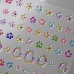 May include: A sheet of colorful flower stickers with a 3D effect. The stickers are arranged in rows and columns on a clear background. The flowers are in various colors, including pink, purple, blue, green, and yellow.